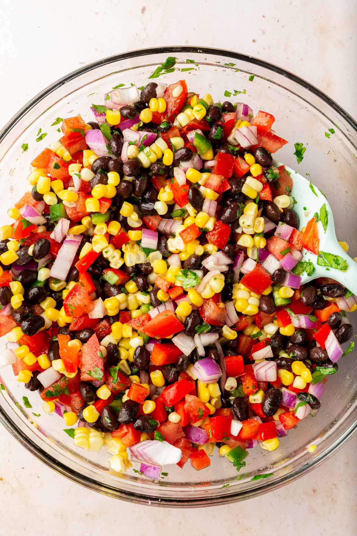 A glass mixing bowl with a corn, black bean, red bell pepper salsa with a blue spatula.