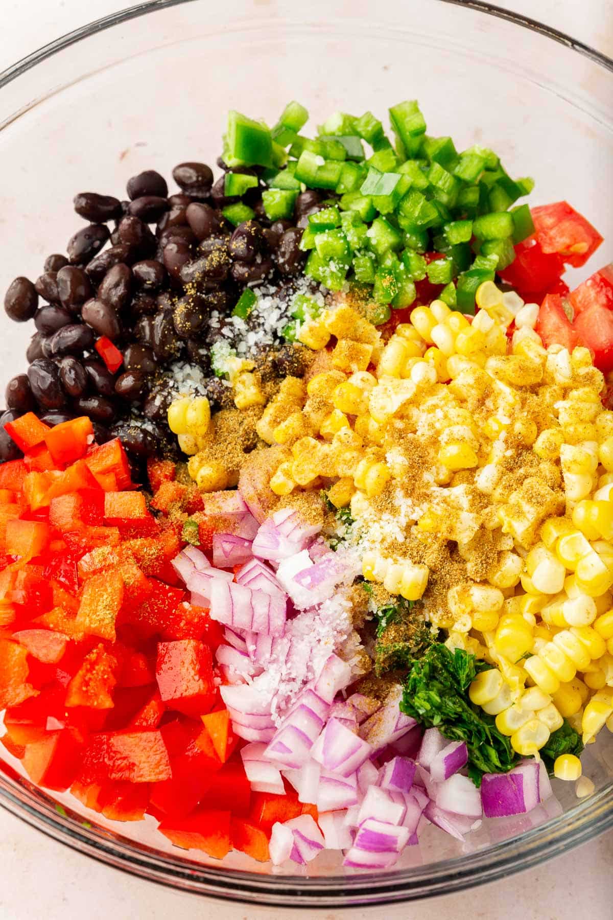 A glass mixing bowl with black beans, jalapeño, tomatoes, red bell pepper, corn, cilantro, cumin and salt before mixing together.