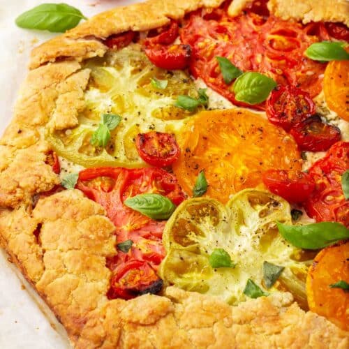 A close up of a gluten-free tomato galette on a piece of parchment paper with cherry tomatoes and basil to the side.