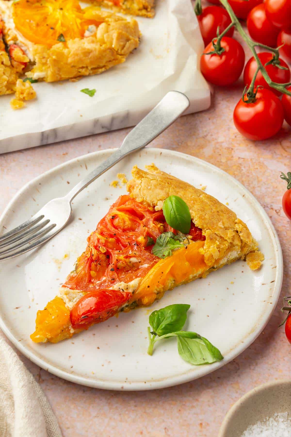 A slice of gluten-free tomato galette on an appetizer plate with basil and a fork with cherry tomatoes on the vine in the background.