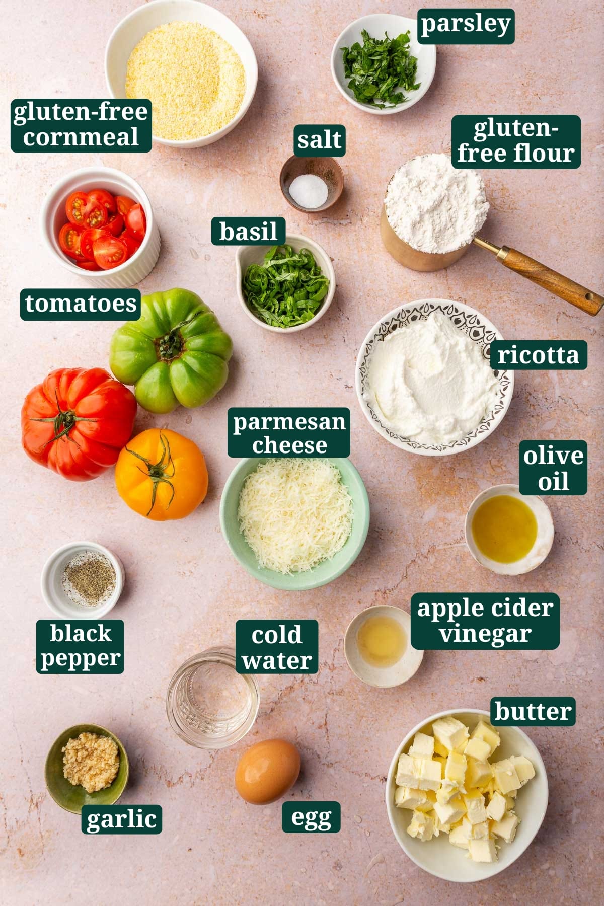 An overhead view of ingredients in small bowls to make a gluten-free tomato galette, including cornmeal, basil, salt, gluten-free flour, parsley, cherry tomatoes, heirloom tomatoes, ricotta cheese, parmesan cheese, pepper, water, olive oil, apple cider vinegar, egg, minced garlic and cold butter with text overlays over each ingredient.