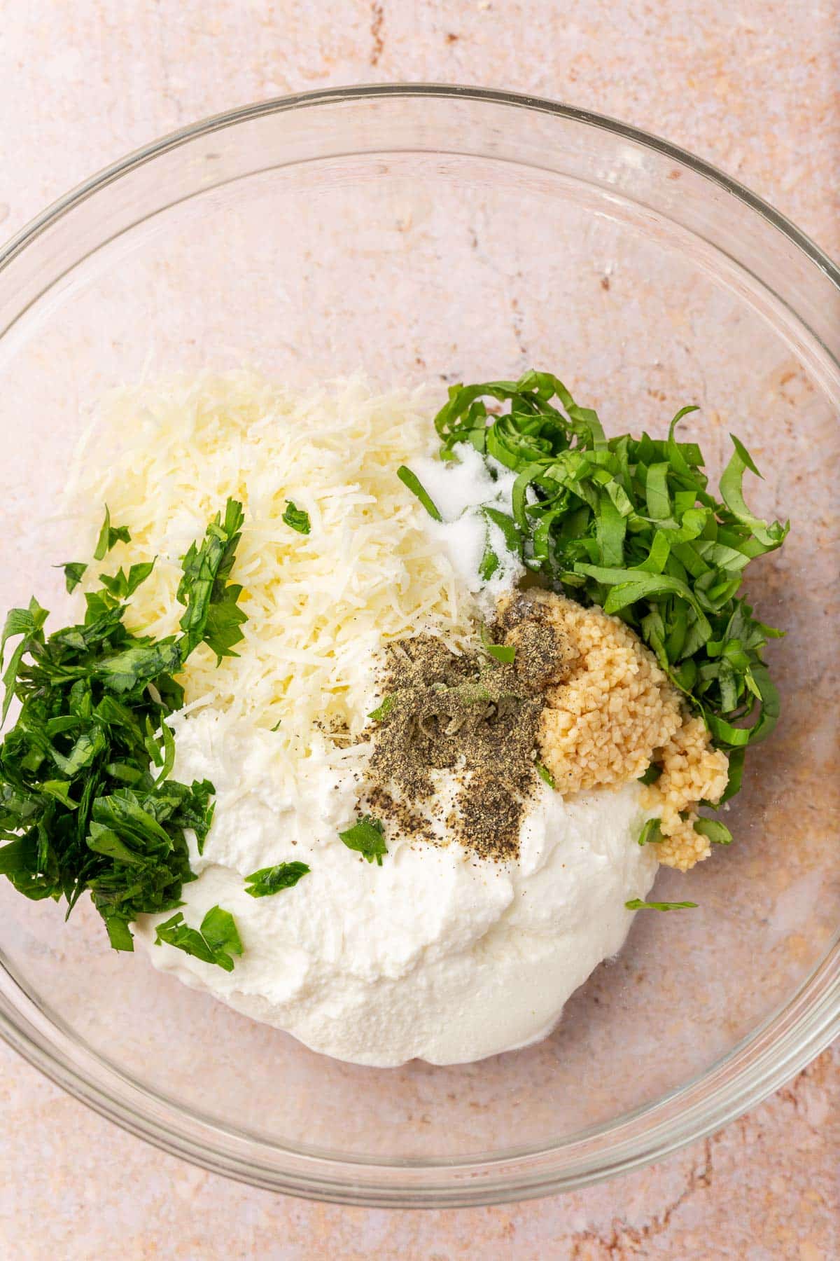 A glass mixing bowl with ricotta cheese, parmesan cheese, parsley, basil, minced garlic, salt, and pepper before mixing together.