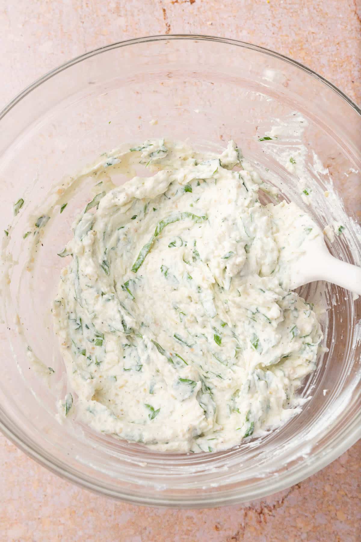 A glass mixing bowl of ricotta cheese and fresh herbs that have been mixed together with a rubber spatula.