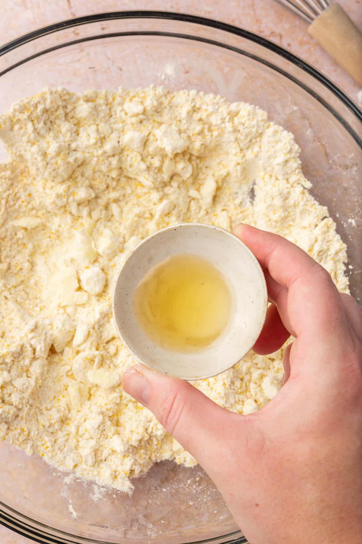 A hand holding a small bowl of apple cider vinegar over a gluten-free flour and butter mixture.