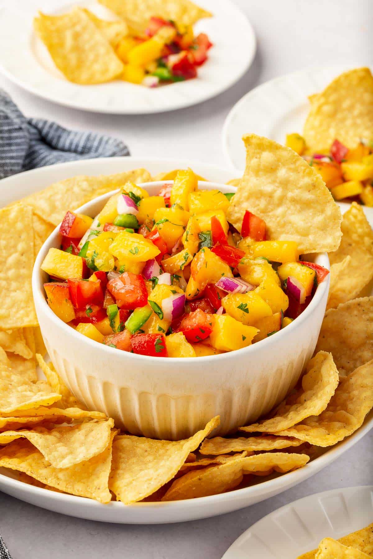A bowl of peach tomato salsa with mango in a large bowl of tortilla chips with appetizer plates of chips and salsa in the background.