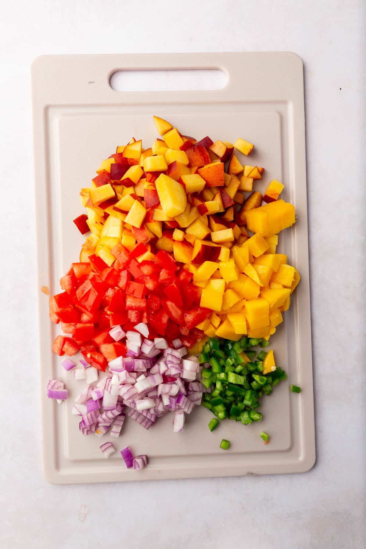 A cutting board with diced peaches, tomatoes, mango, red onion and jalapeño on it.