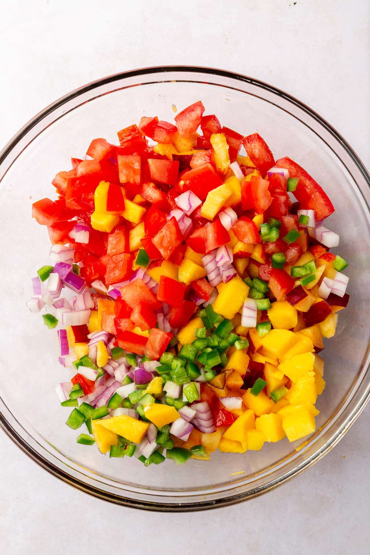 A glass mixing bowl with diced tomatoes, mango, peaches, red onion and jalapeños in it before mixing together.