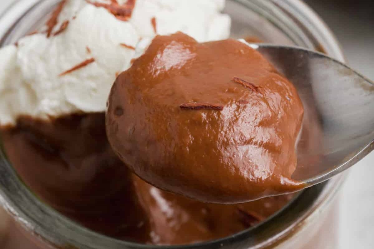 A close-up of a spoonful of creamy chocolate pudding with chocolate shavings, held above a jar topped with whipped cream.