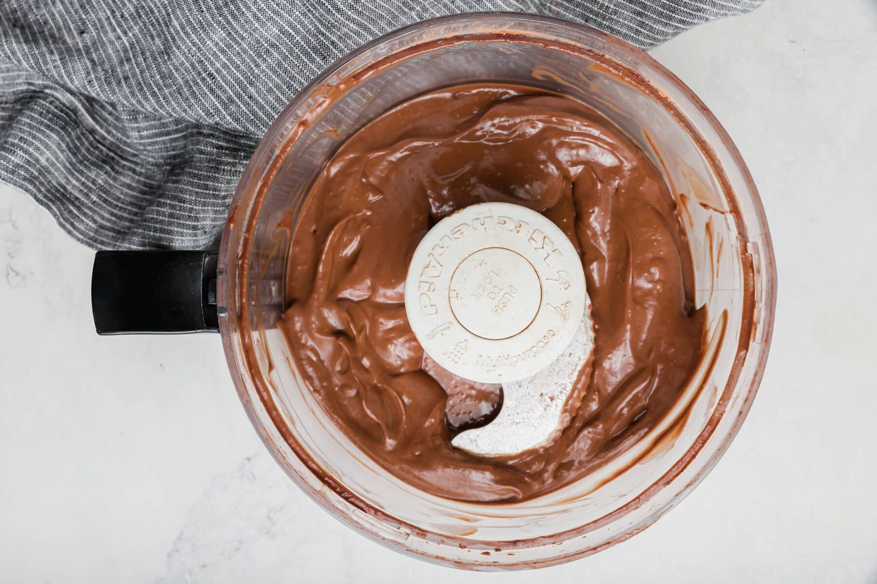 A food processor filled with smooth, creamy chocolate mixture, viewed from above. The machine sits on a light marble surface next to a gray striped cloth.