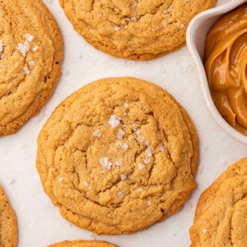 GF peanut butter cookies with flaky sea salt on a piece of parchment paper with a bowl of peanut butter peaking in.