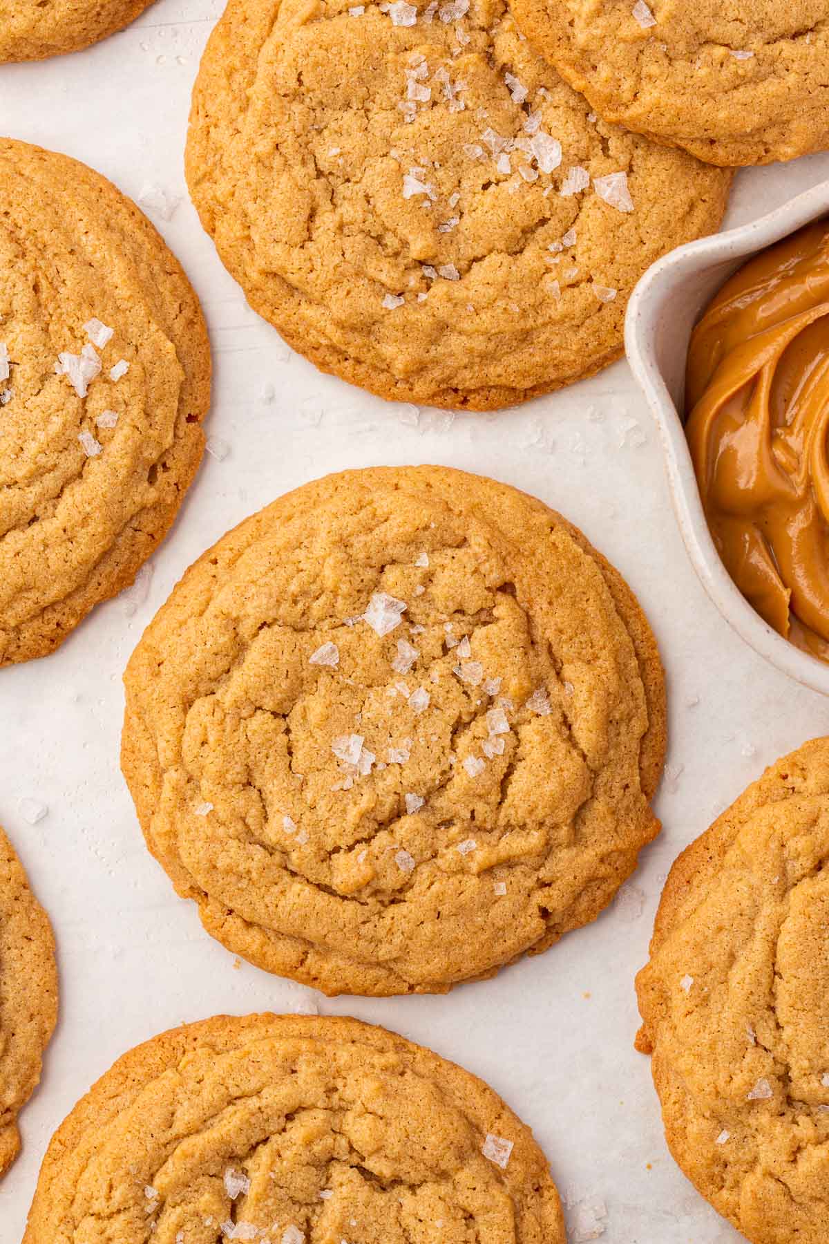 GF peanut butter cookies with flaky sea salt on a piece of parchment paper with a bowl of peanut butter peaking in.