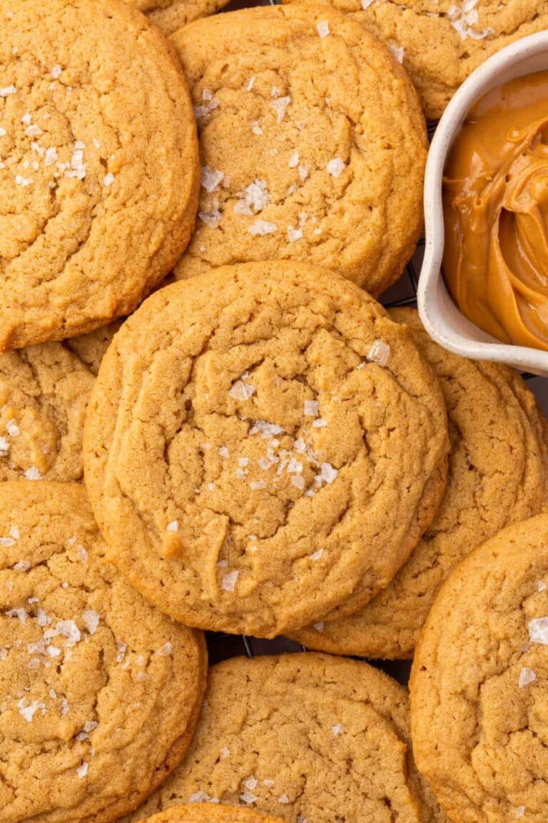 A pile of gluten-free peanut butter cookies topped with flaky sea salt and a bowl of peanut butter on the side.