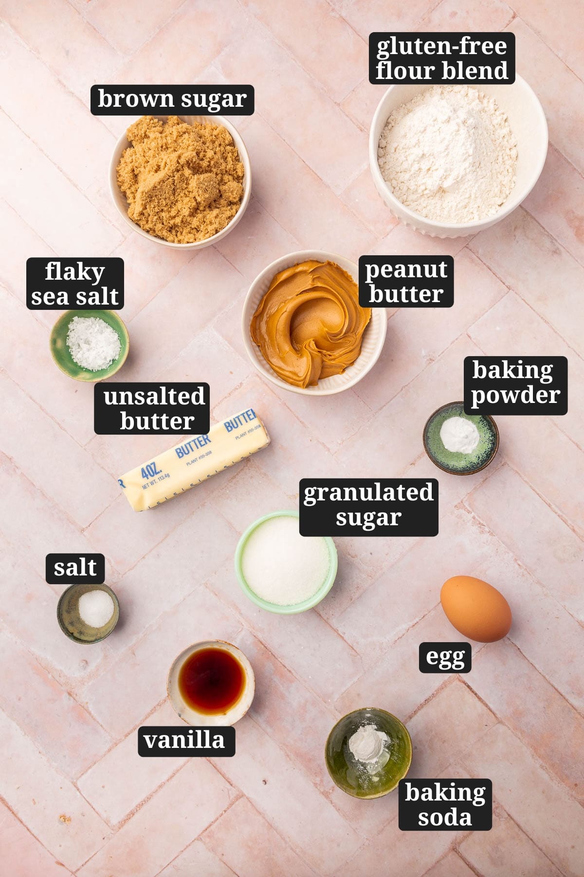 Ingredients in small bowls to make gluten-free peanut butter cookies, including brown sugar, gluten-free flour, flaky sea salt, peanut butter, butter, baking soda, baking powder, granulated sugar, vanilla, egg, and salt with text overlays over each ingredient.