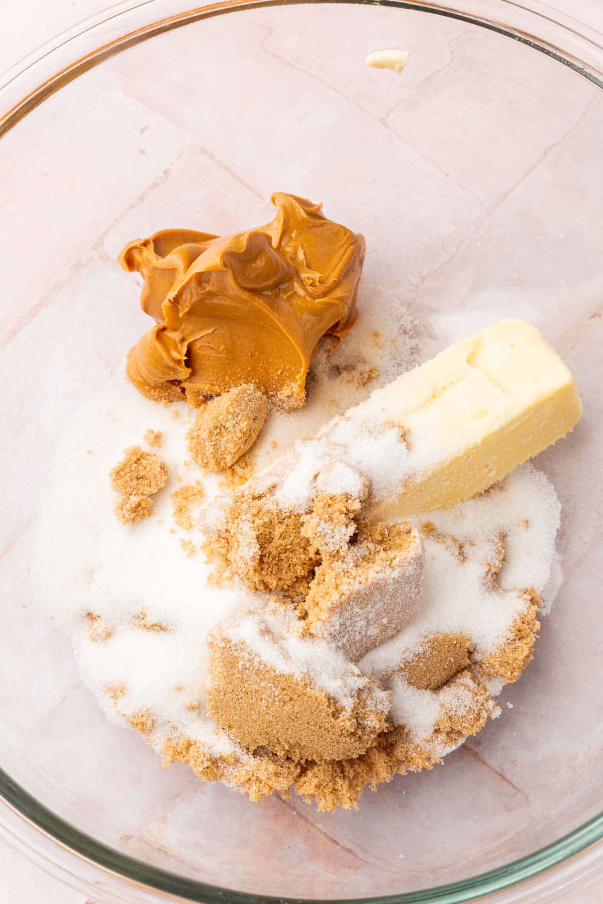 A glass mixing bowl with peanut butter, a stick of butter, granulated sugar and brown sugar in it before mixing together.
