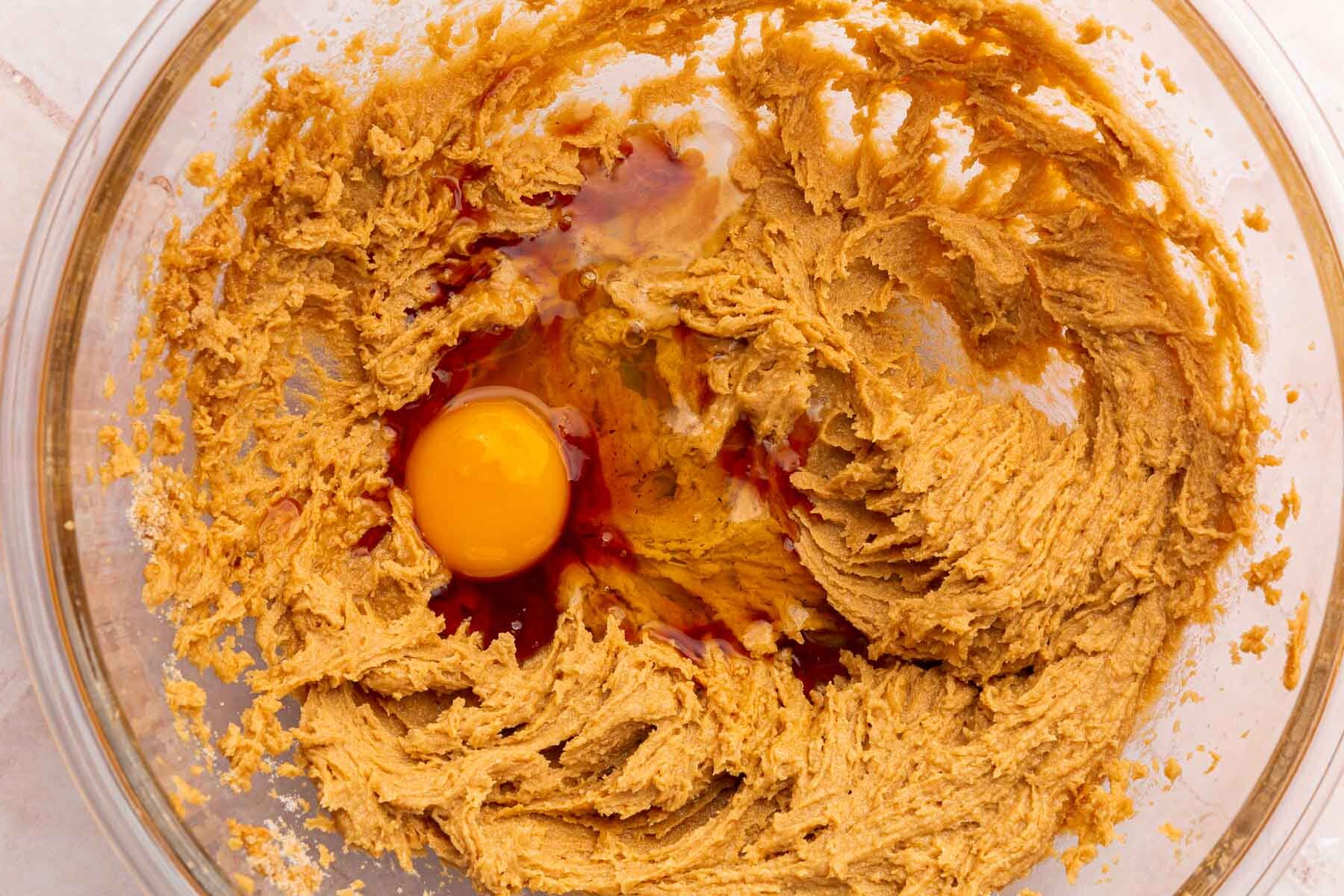 A glass mixing bowl with a peanut butter sugar mixture topped with a raw egg and vanilla extract before mixing together.
