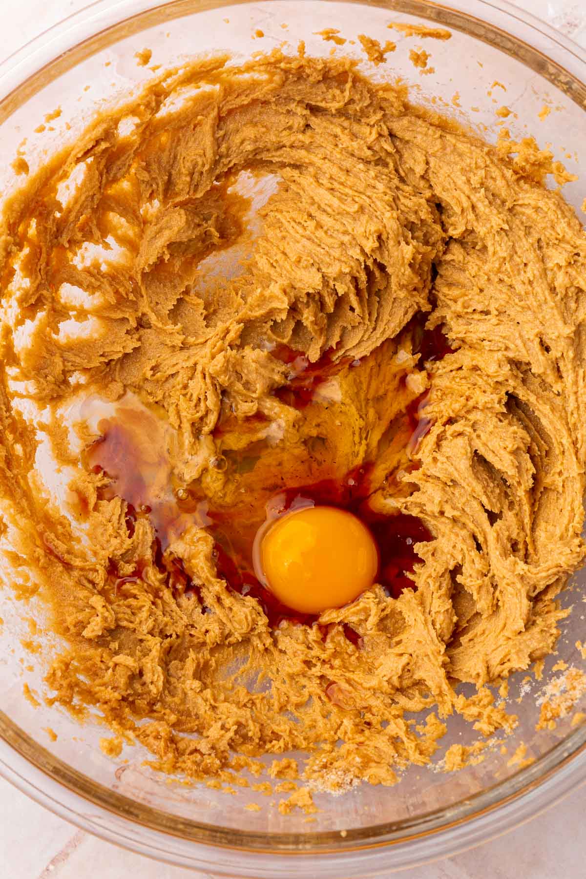 A glass mixing bowl with a peanut butter sugar mixture topped with a raw egg and vanilla extract before mixing together.