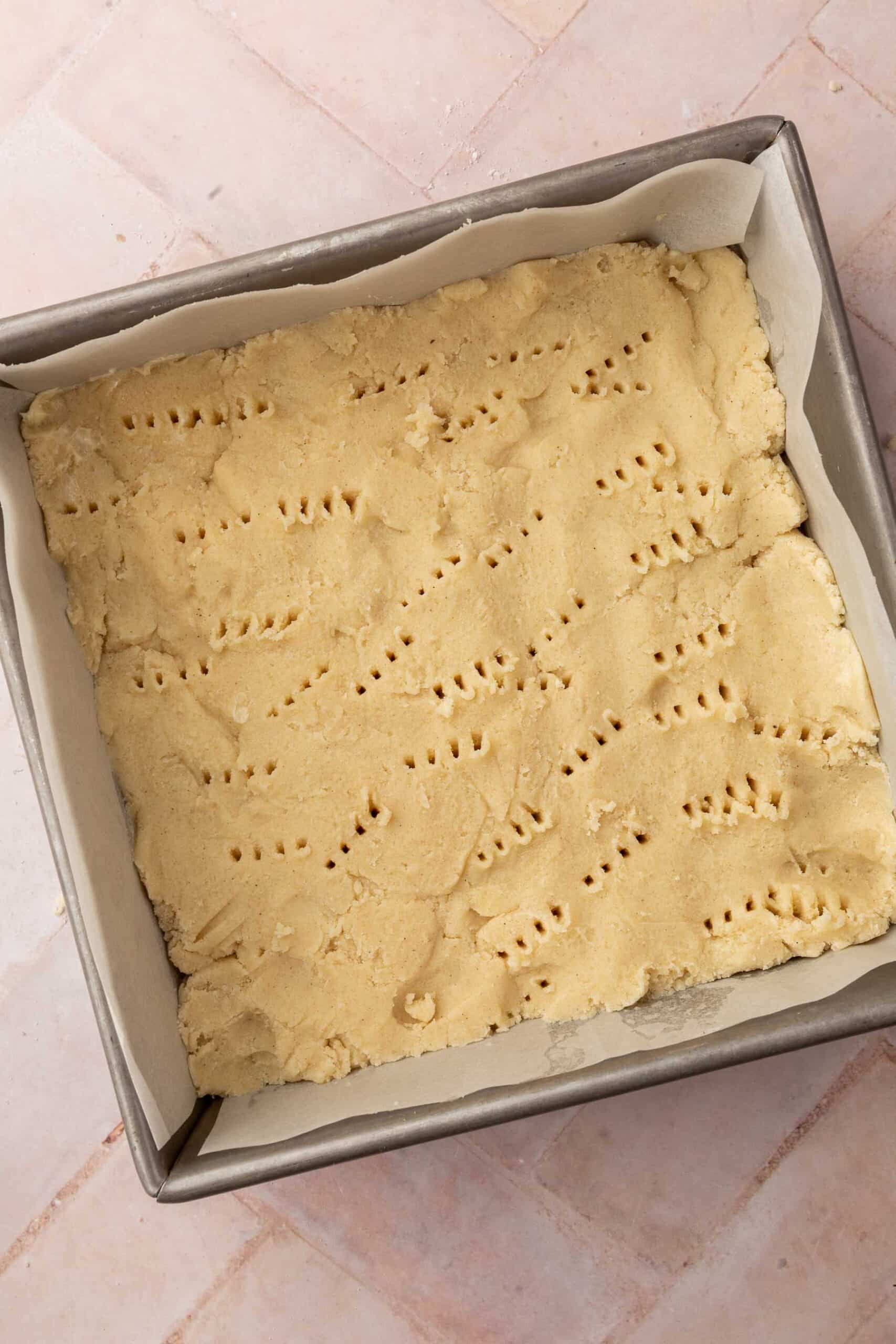 A square baking pan lined with parchment paper with gluten-free shortbread dough in it with fork holes poked all over.