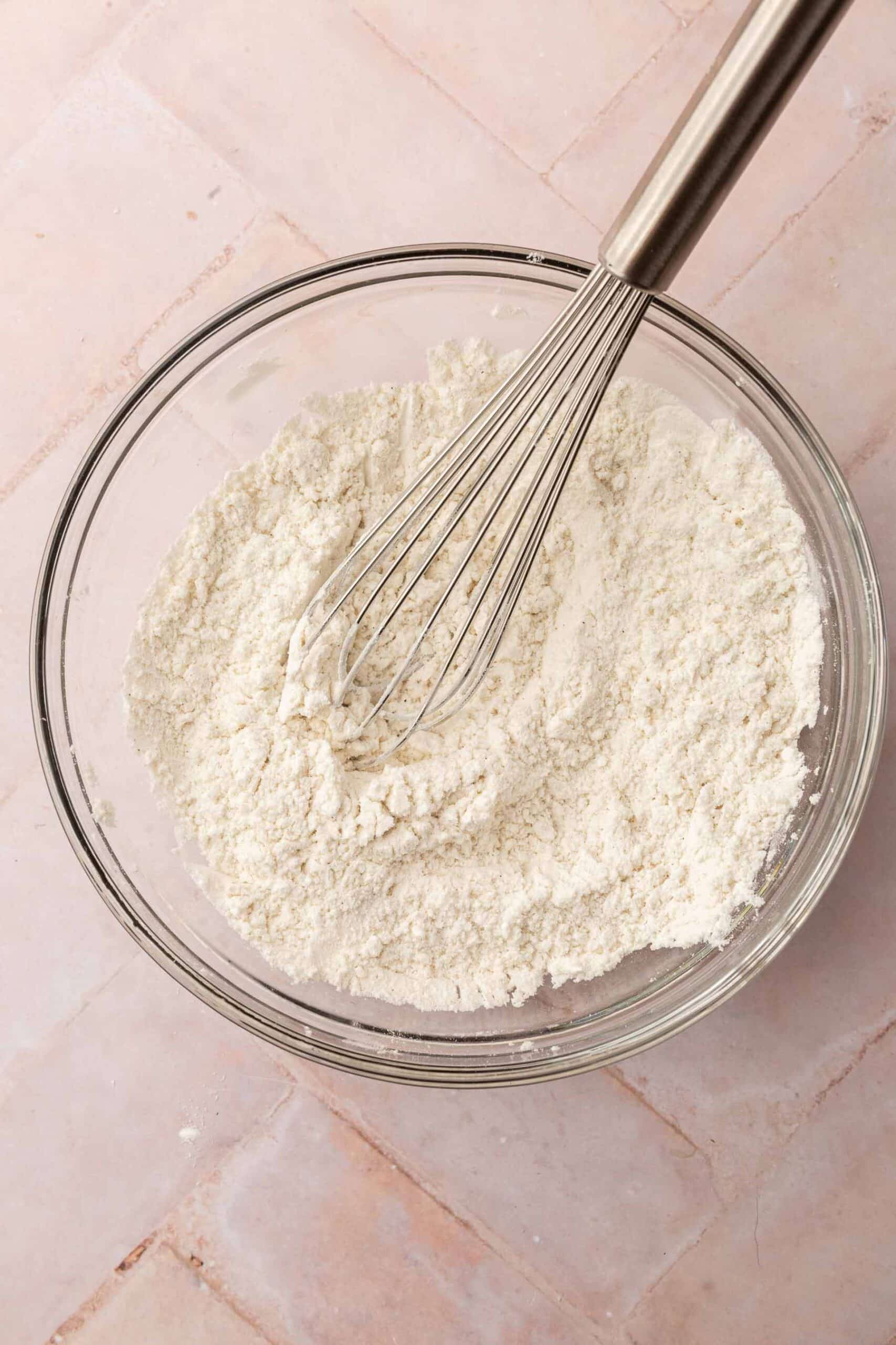 A glass mixing bowl with gluten-free flour, cornstarch and salt in it with a metal wire whisk.