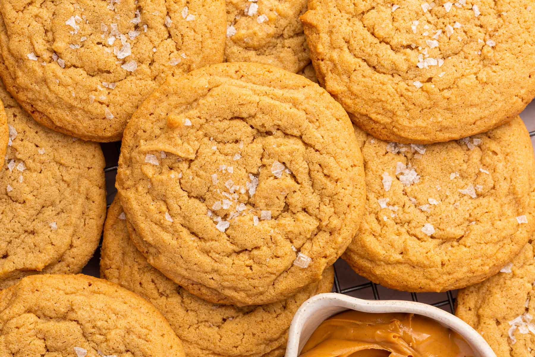 A pile of gluten-free peanut butter cookies topped with flaky sea salt and a bowl of peanut butter on the side.
