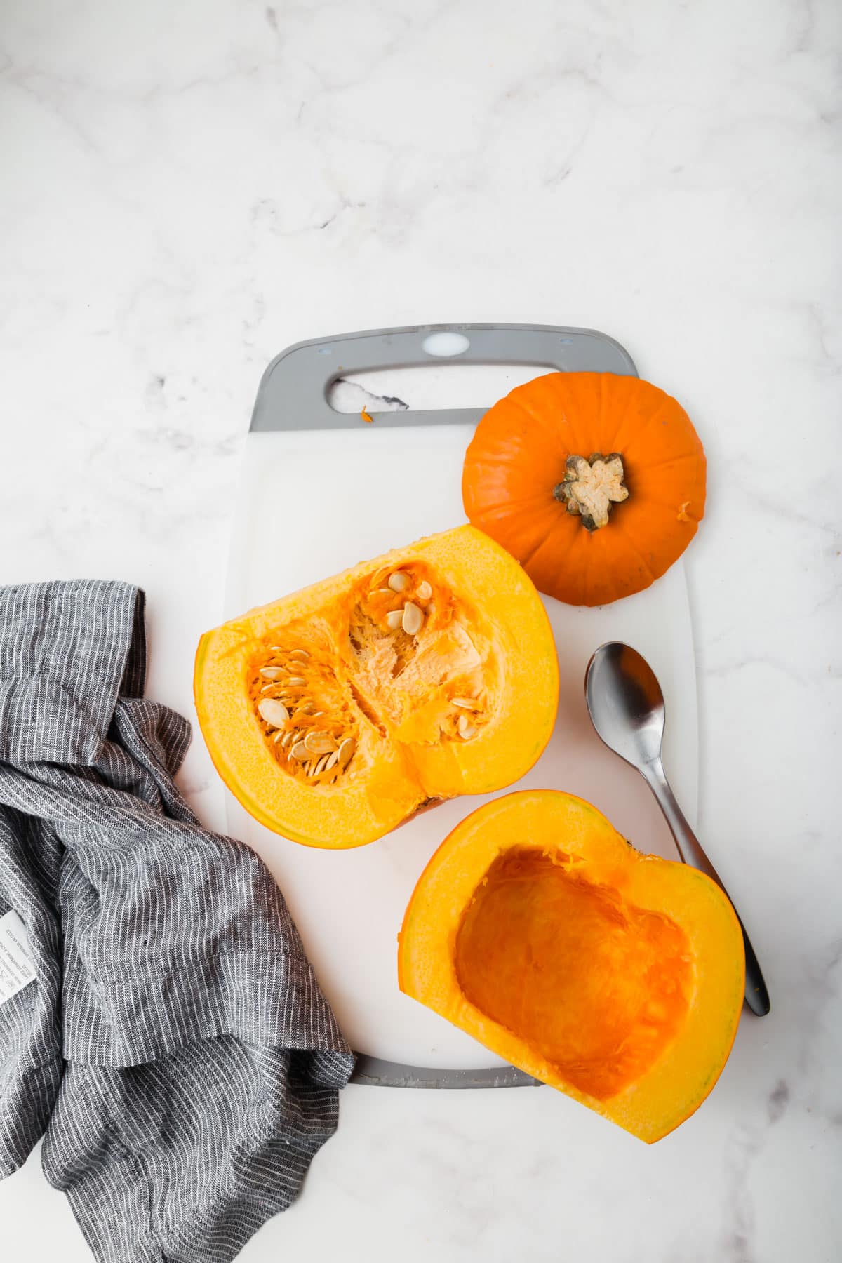 A sugar pumpkin sliced in half on a cutting board with a spoon next to it to scoop out the seeds.