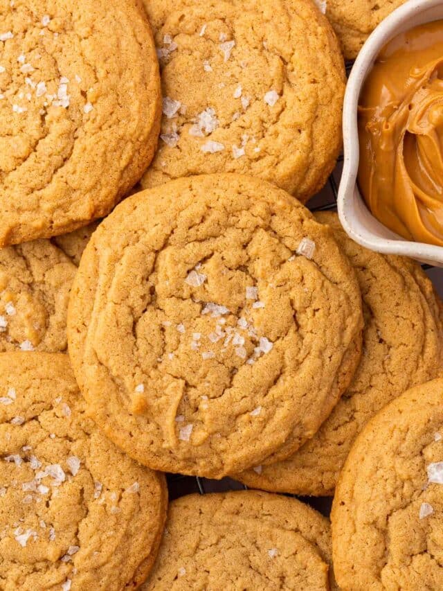 A pile of gluten-free peanut butter cookies topped with flaky sea salt and a bowl of peanut butter on the side.
