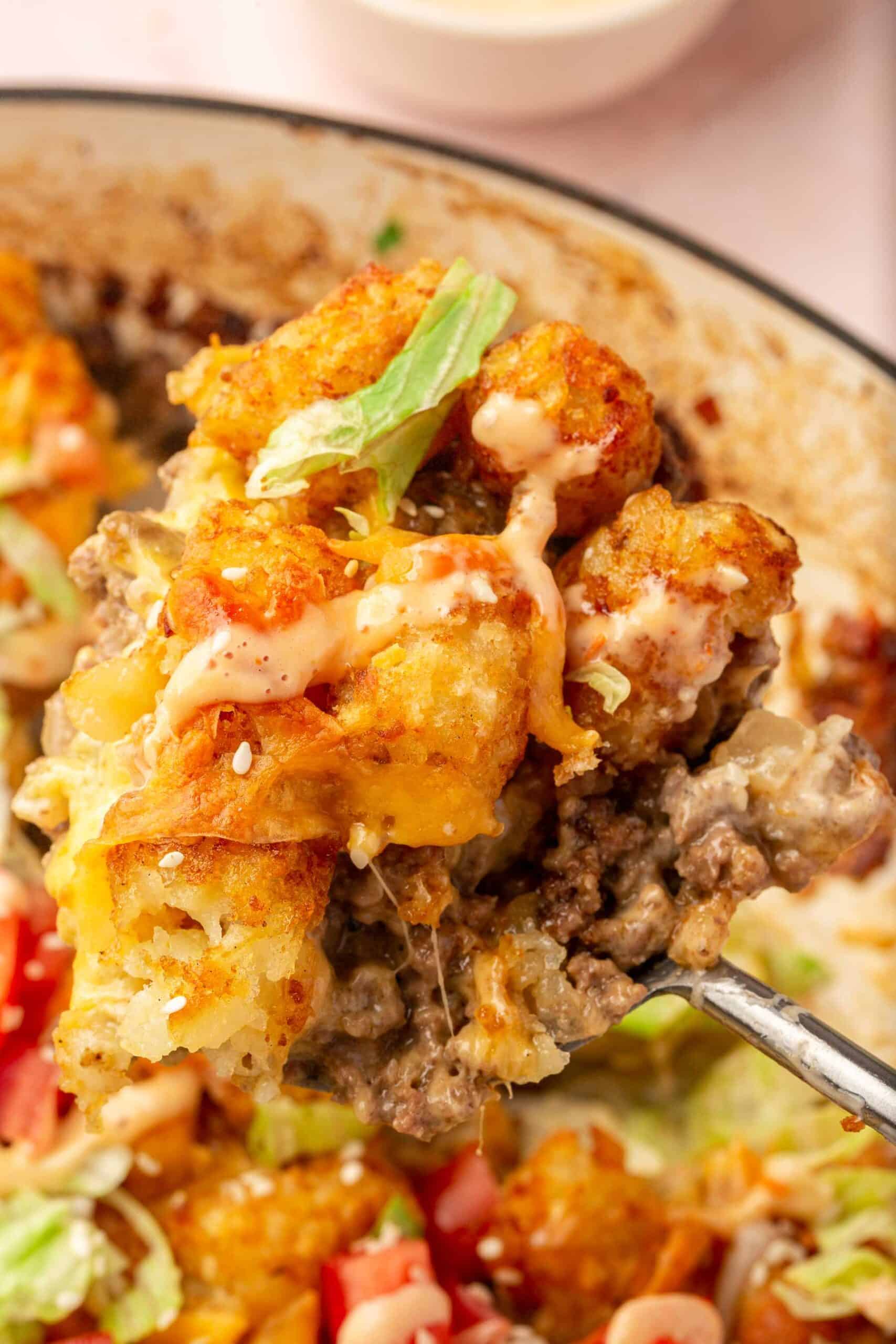 A large serving spoon scooping a portion of big mac casserole with tater tots out of the casserole dish.