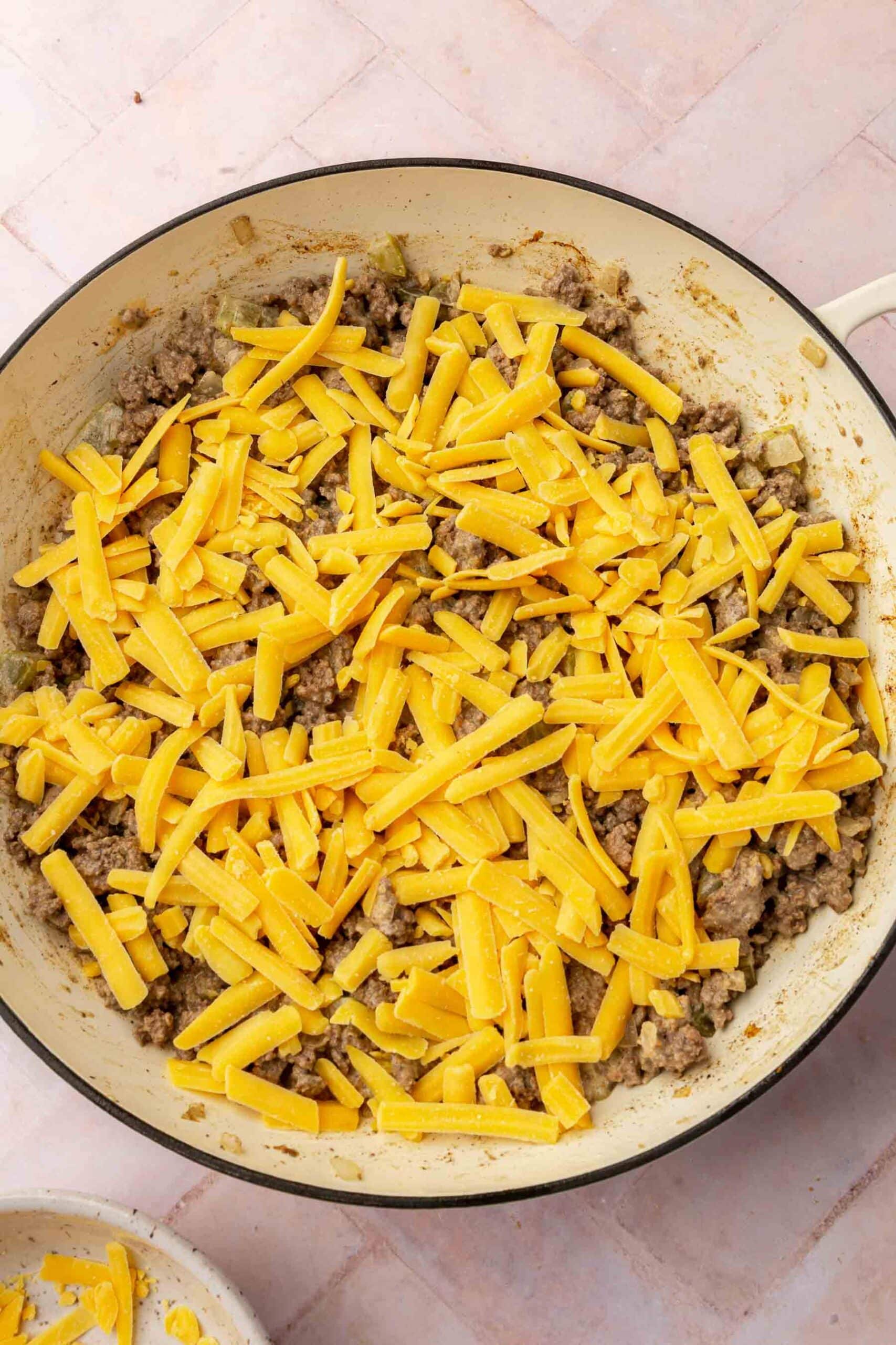 A braising pan with a ground beef mixture topped with shredded cheddar cheese before baking in the oven.