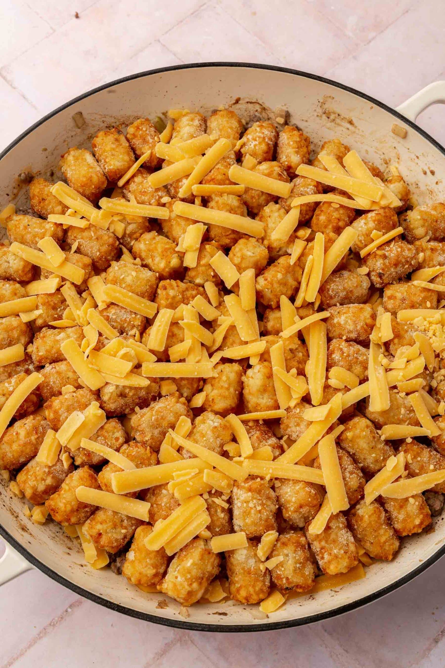 Tater tots topped with shredded cheese in a round white braising pan before placing in the oven.