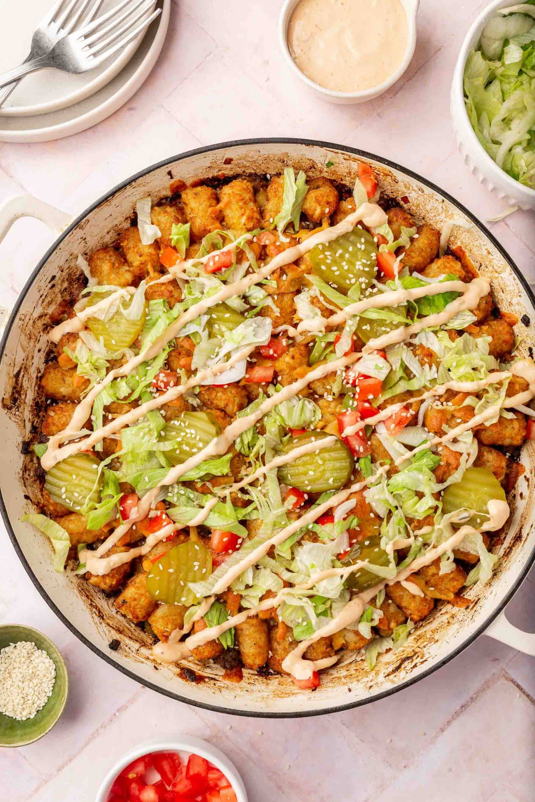 Big mac casserole in a white braising pan topped with lettuce, tomatoes, pickles, big mac sauce and sesame seeds.