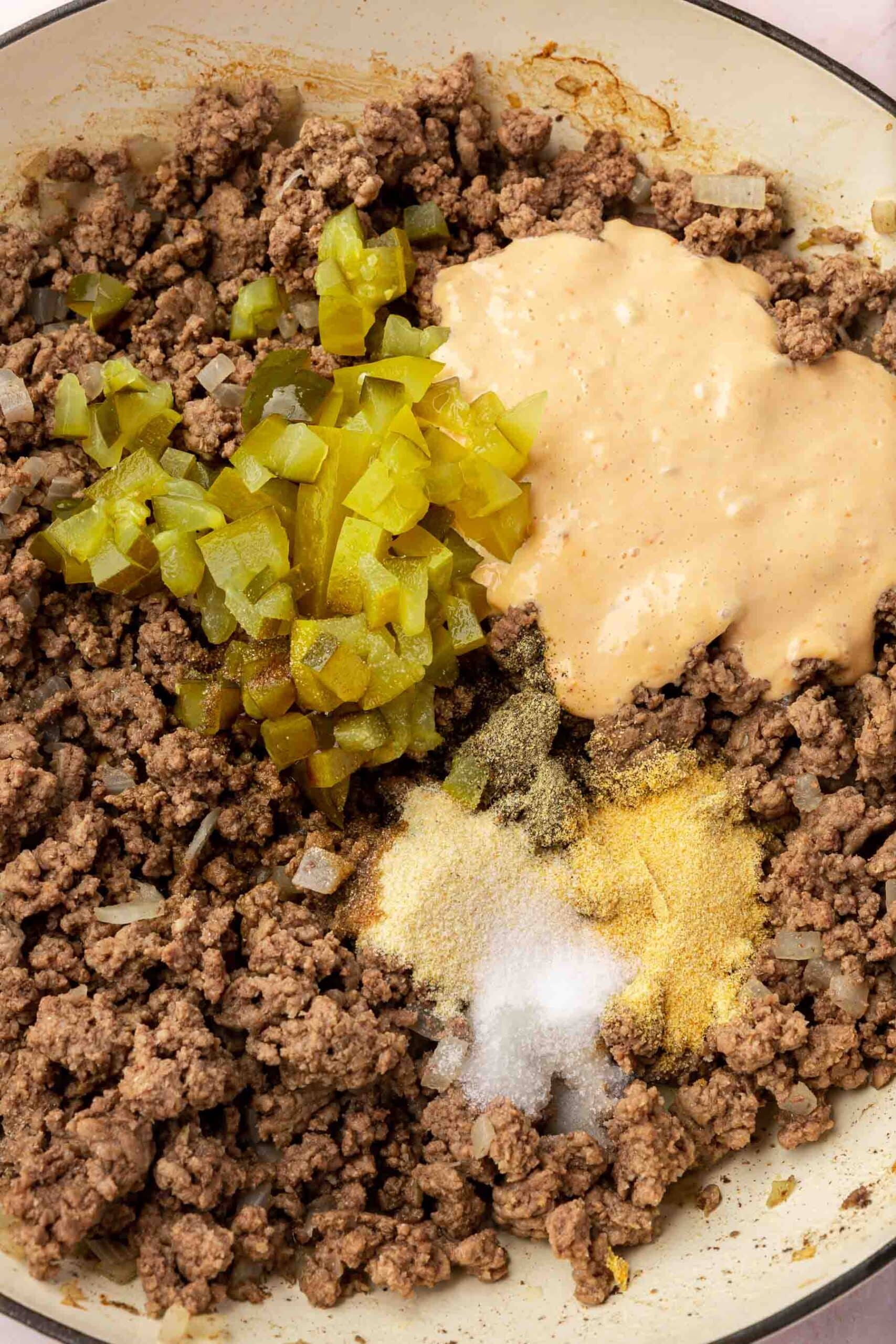 Cooked ground beef in a braising pan topped with diced pickles, garlic powder, salt, onion powder and thousand island dressing before mixing together.