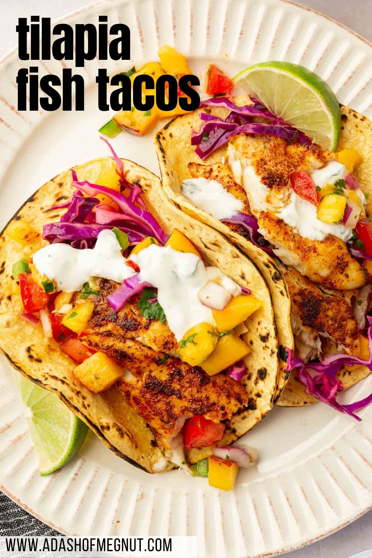 Two tilapia fish tacos topped with fruit salsa, cabbage slaw, sour cream and lime wedges on a plate.