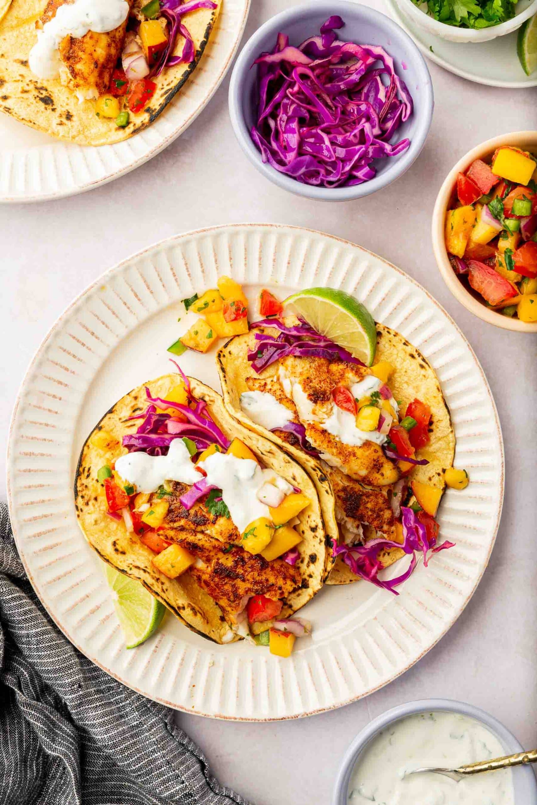 An overhead view of two fish tacos on charred corn tortillas topped with sour cream, fruit salsa, and red cabbage with bowls of slaw, fruit salsa, and crema to the side.