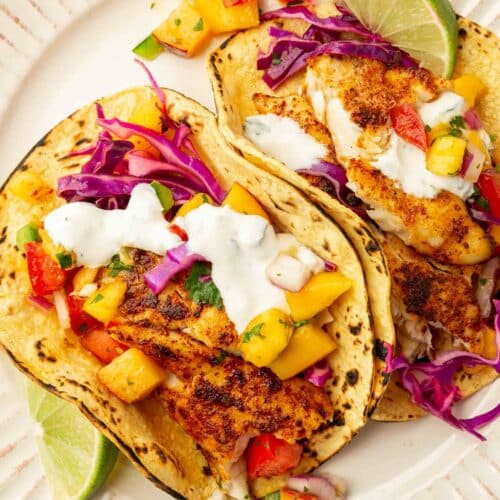 An overhead view of two tilapia fish tacos topped with lime crema, red cabbage slaw, and peach mango salsa on charred corn tortillas.