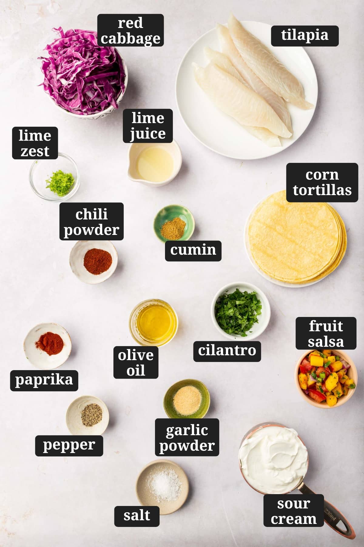 Ingredients in small bowls to make tilapia fish tacos, including red cabbage, raw tilapia filets, lime zest, lime juice, cumin, paprika, chili powder, olive oil, cilantro, corn tortillas, pepper, garlic powder, salt, sour cream, and fruit salsa with text overlays over each ingredient.