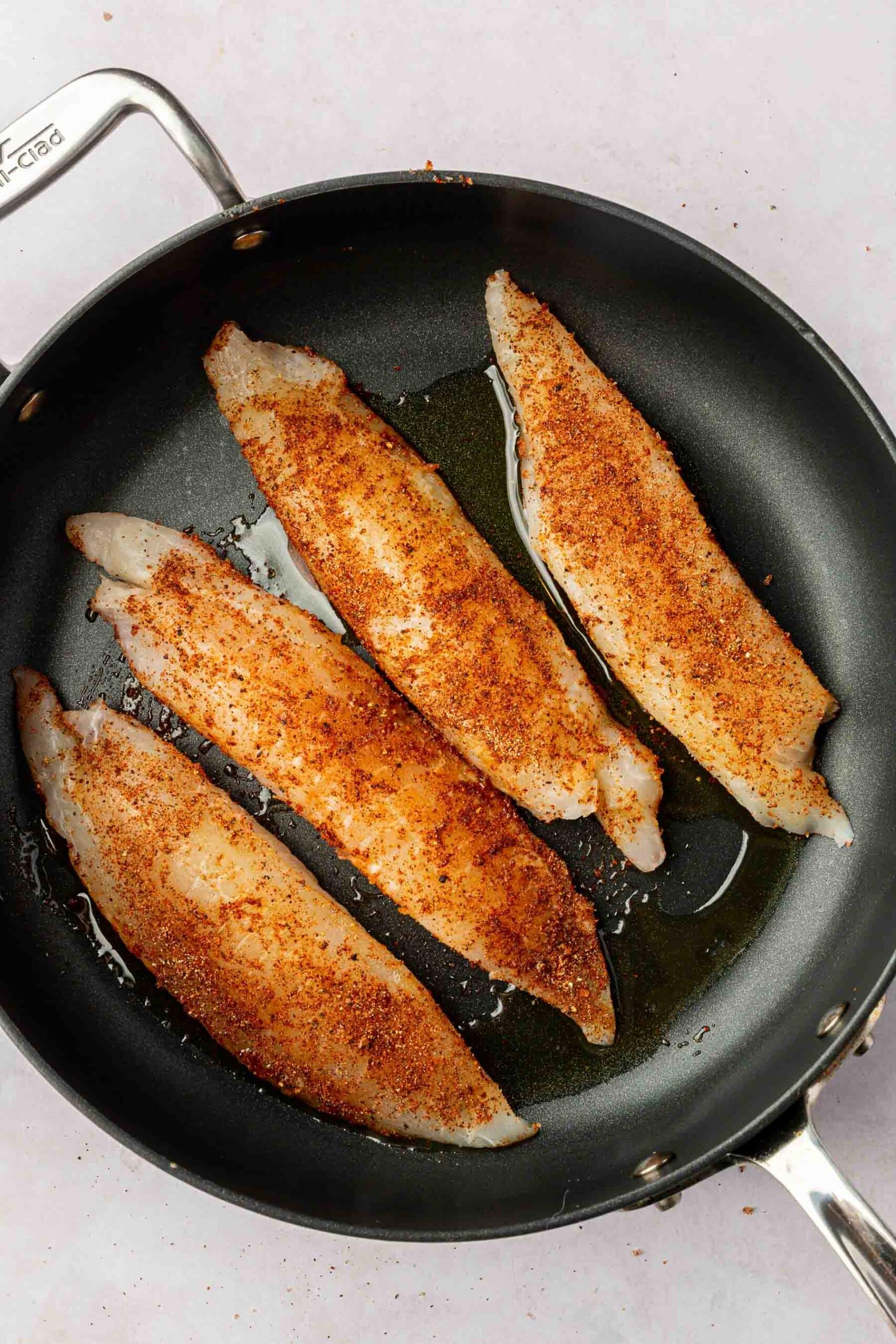 Four raw tilapia loin filets covered in seasoning in a non-stick skillet with olive oil.
