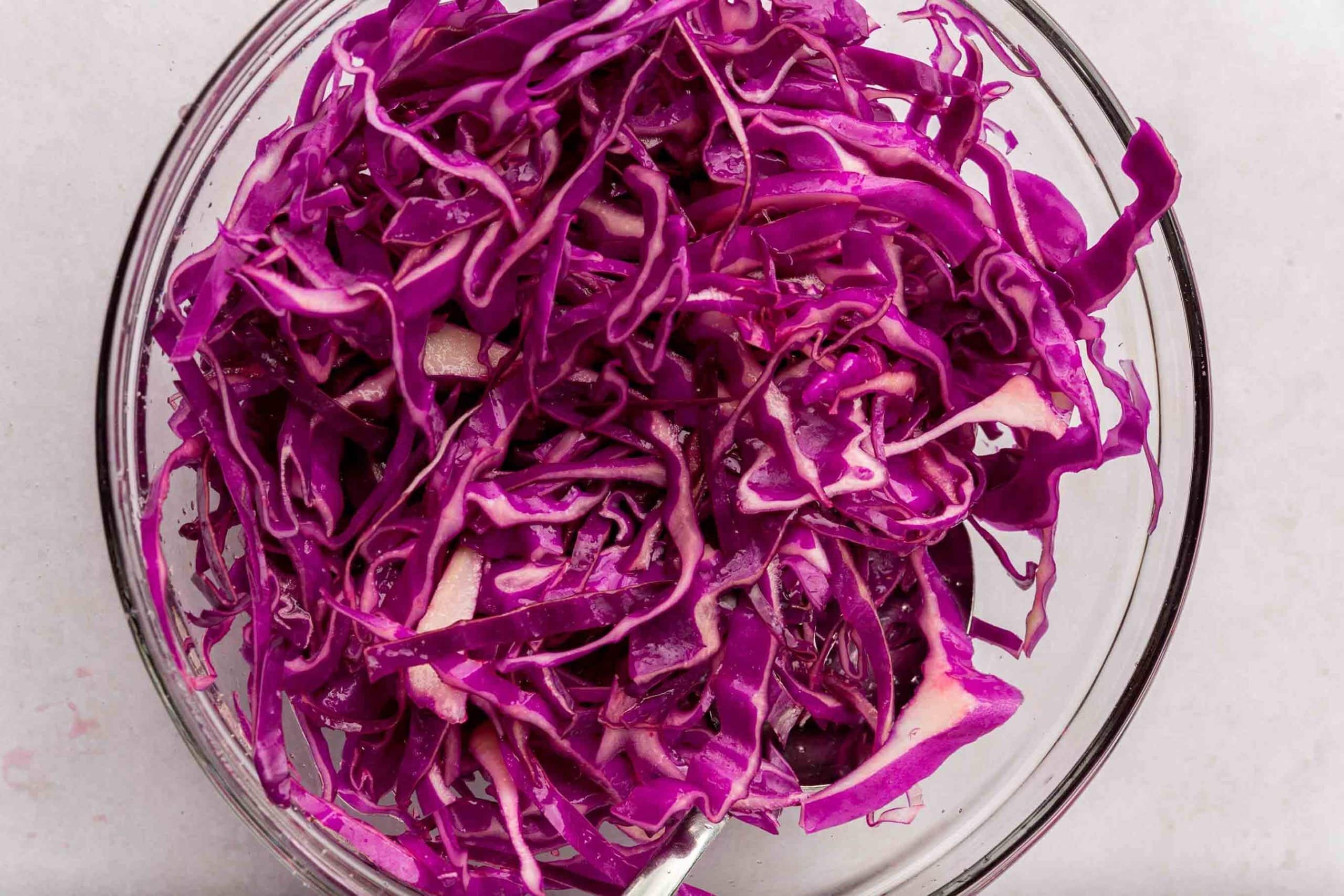 A glass mixing bowl with shredded red cabbage mixed with salt and lime juice.