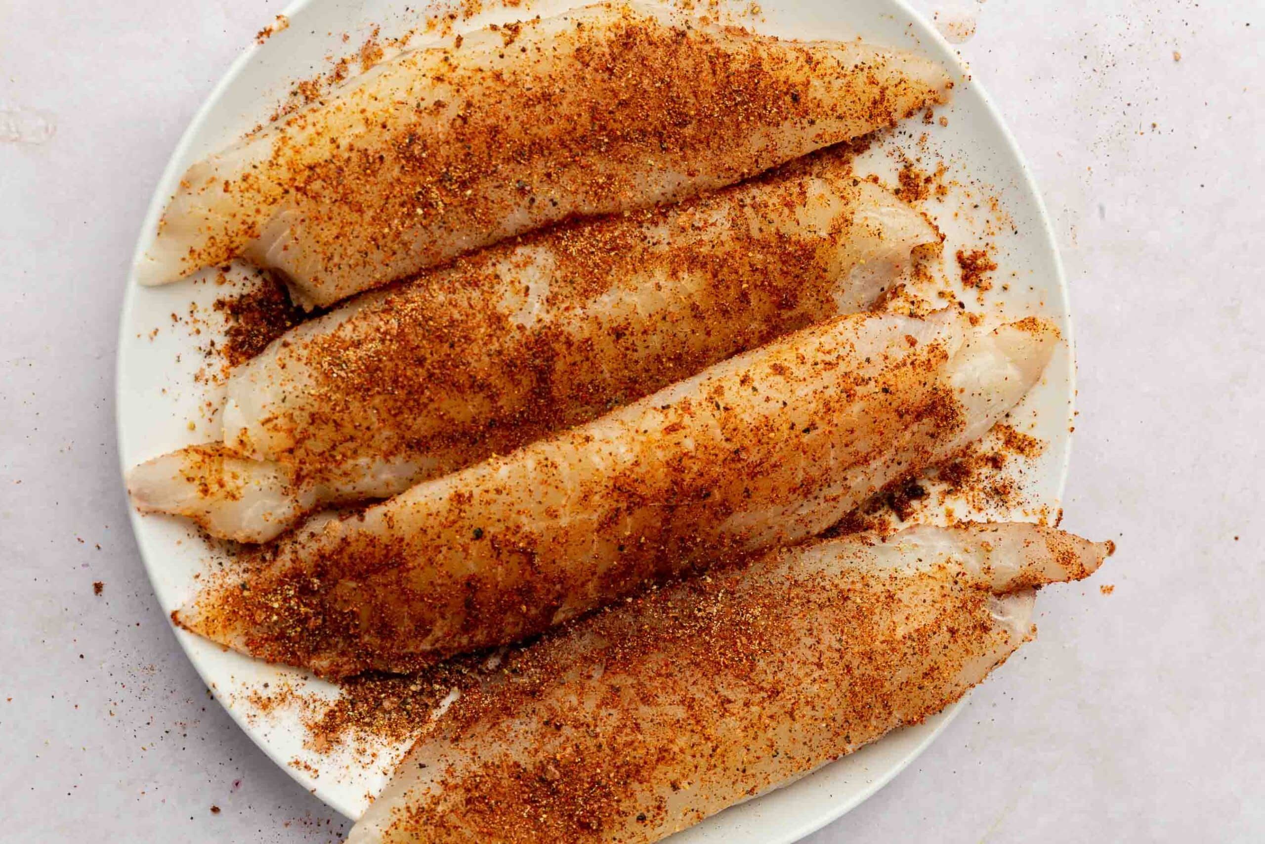 A white plate topped with four tilapia loin filets that have been covered with taco seasoning mix.
