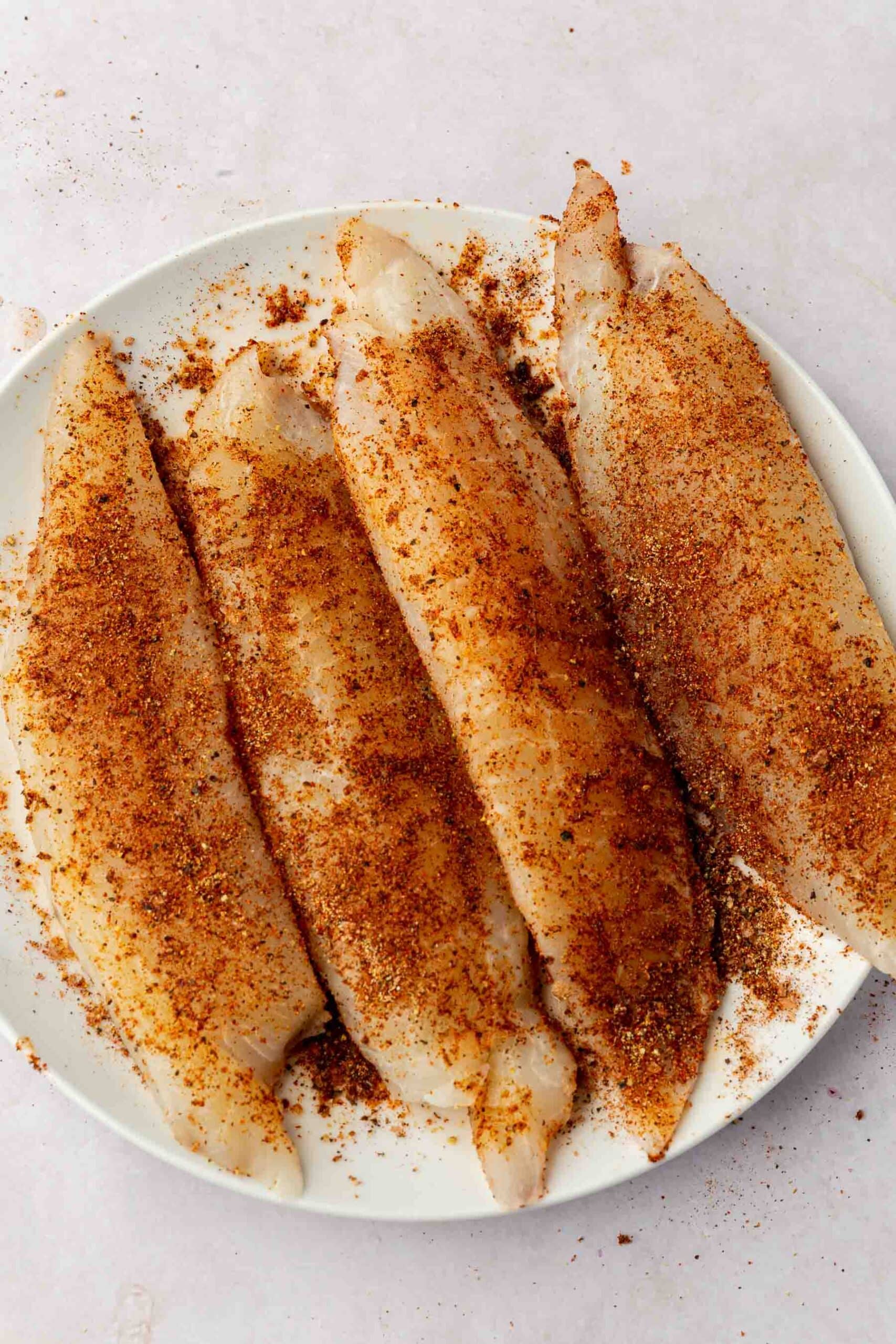 A white plate topped with four tilapia loin filets that have been covered with taco seasoning mix.