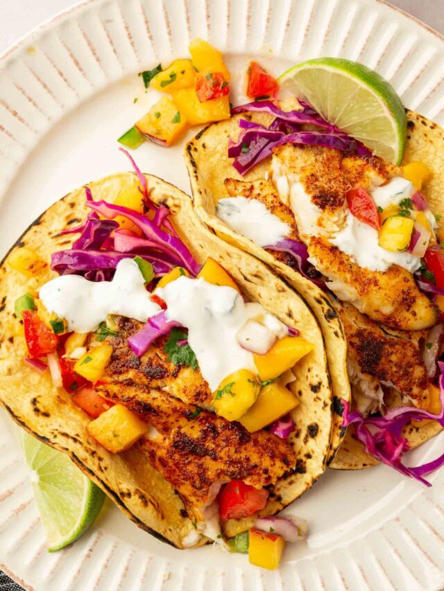 An overhead view of two tilapia fish tacos topped with lime crema, red cabbage slaw, and peach mango salsa on charred corn tortillas.