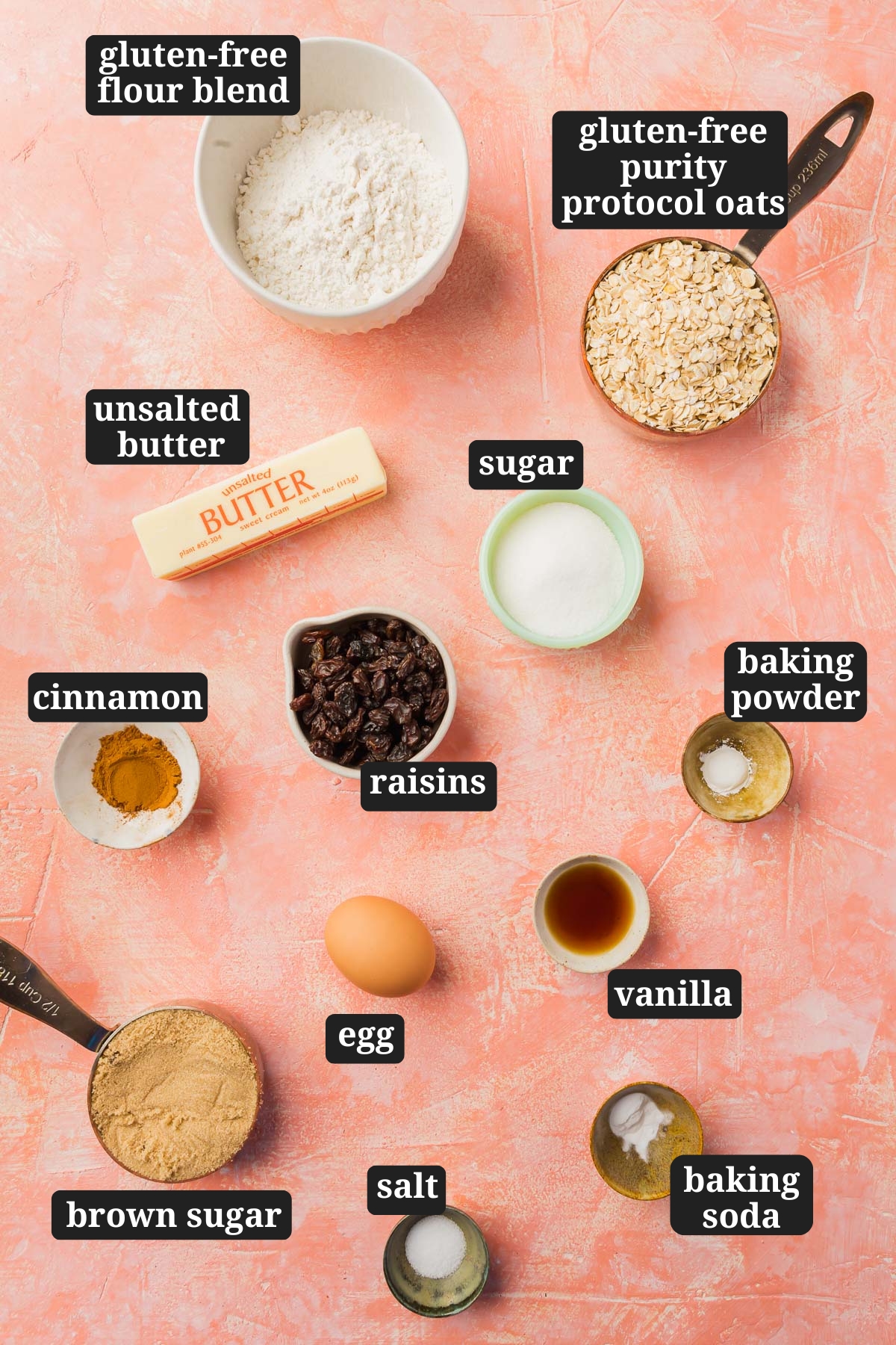 An overhead view of ingredients in small bowls to make gluten free oatmeal raisin cookies, including gluten-free flour blend, purity protocol oats, unsalted butter, sugar, cinnamon, raisins, baking powder, egg, vanilla, brown sugar, salt and baking soda with text overlays over each ingredient.