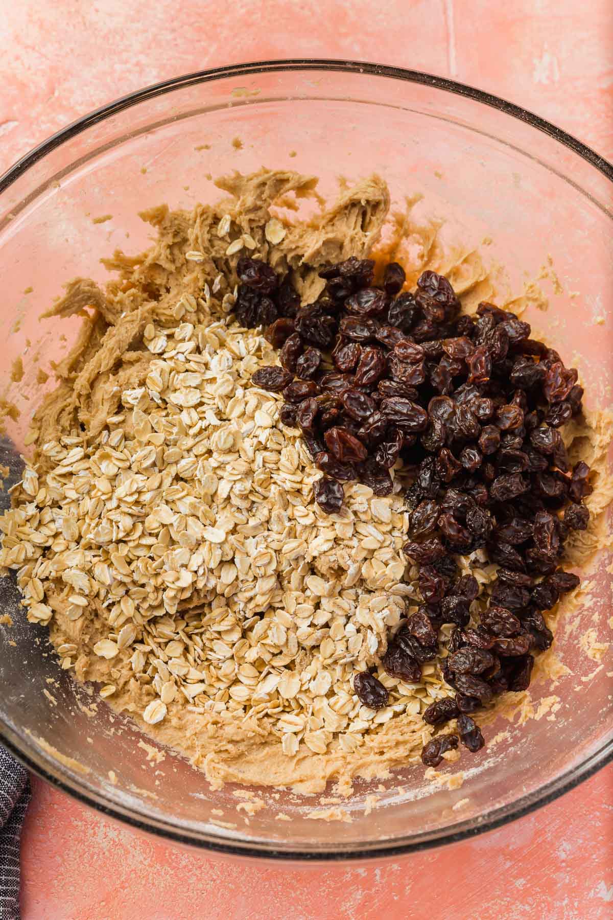 A glass mixing bowl with gluten-free oats and raisins added to gluten free cookie dough on a pink table.