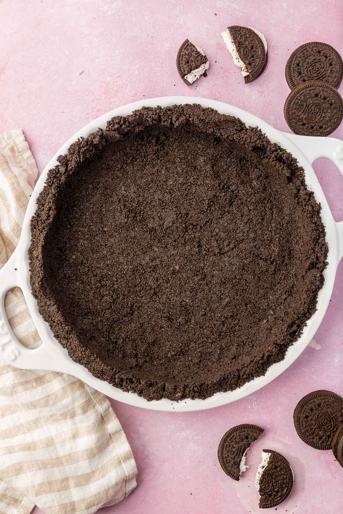 A gluten-free Oreo pie crust pressed into a white tart pan with whole and broken Oreos surrounding it on a pink table.