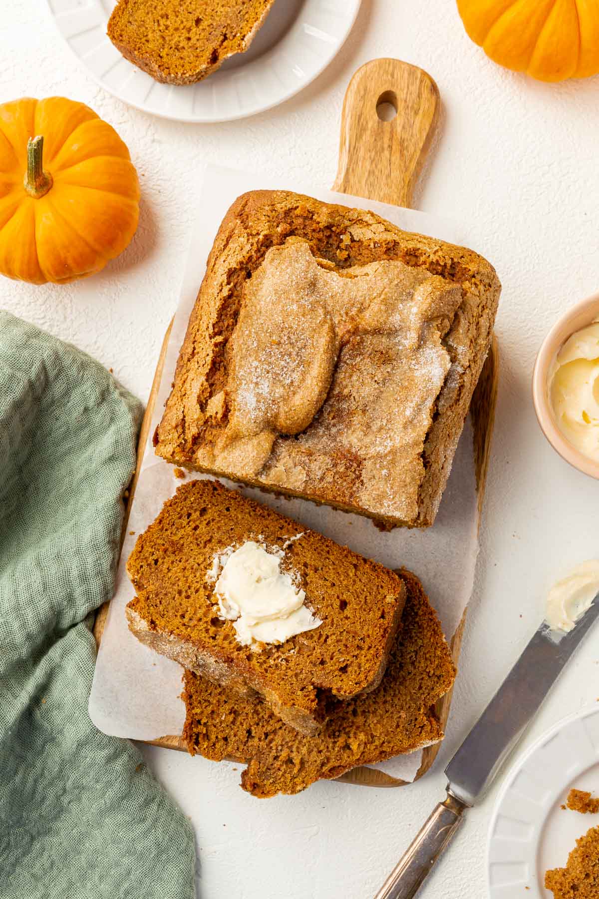 An overhead view of a pumpkin bread loaf that has been partially sliced and topped with a slather of butter with mini pumpkins surrounding the bread.