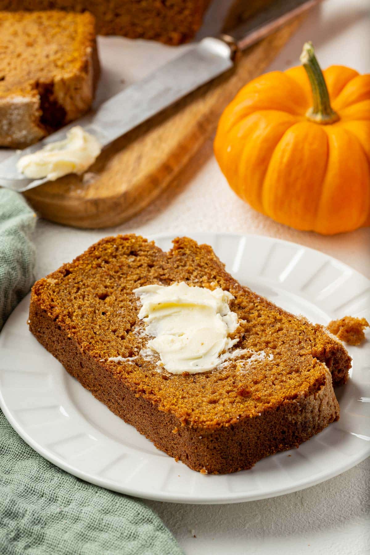 A single slice of gluten free pumpkin bread on a dessert plate slathered with butter with the larger loaf in the background.
