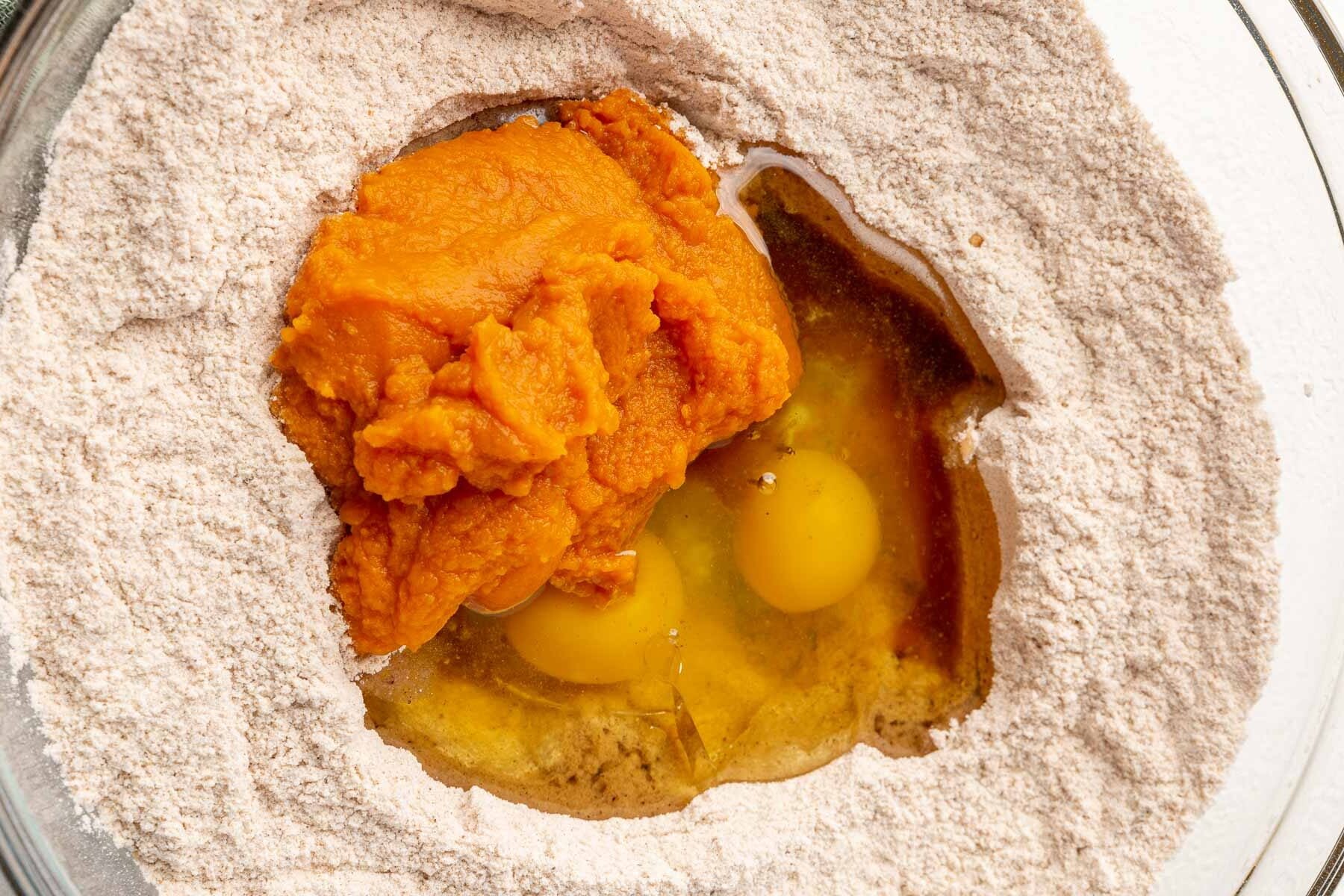 A glass mixing bowl with gluten-free flour topped with vanilla, oil, eggs, and pumpkin puree before mixing together.