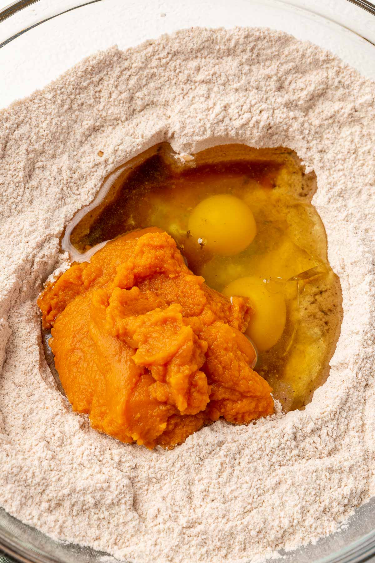 A glass mixing bowl with gluten-free flour topped with vanilla, oil, eggs, and pumpkin puree before mixing together.