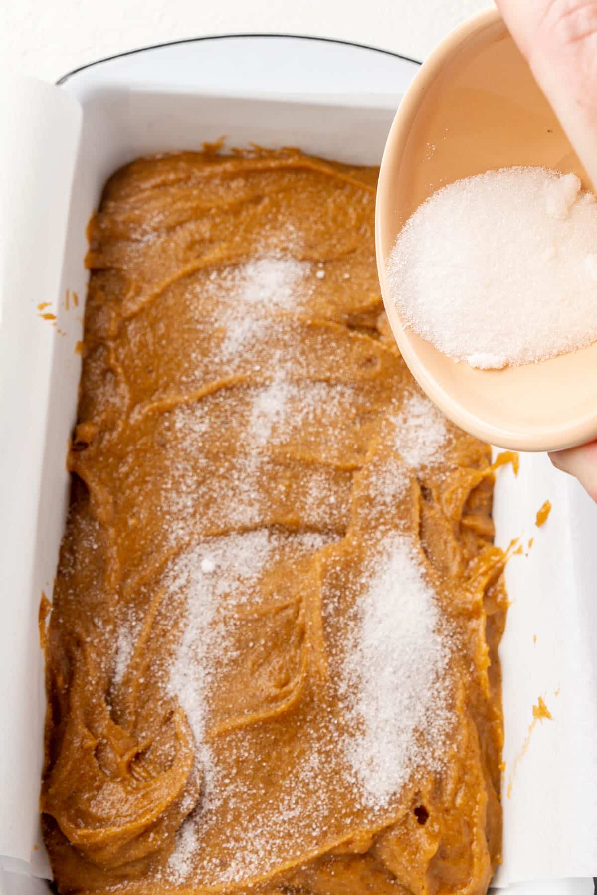 A hand holding a bowl of granulated sugar over a loaf of gluten-free pumpkin bread batter.