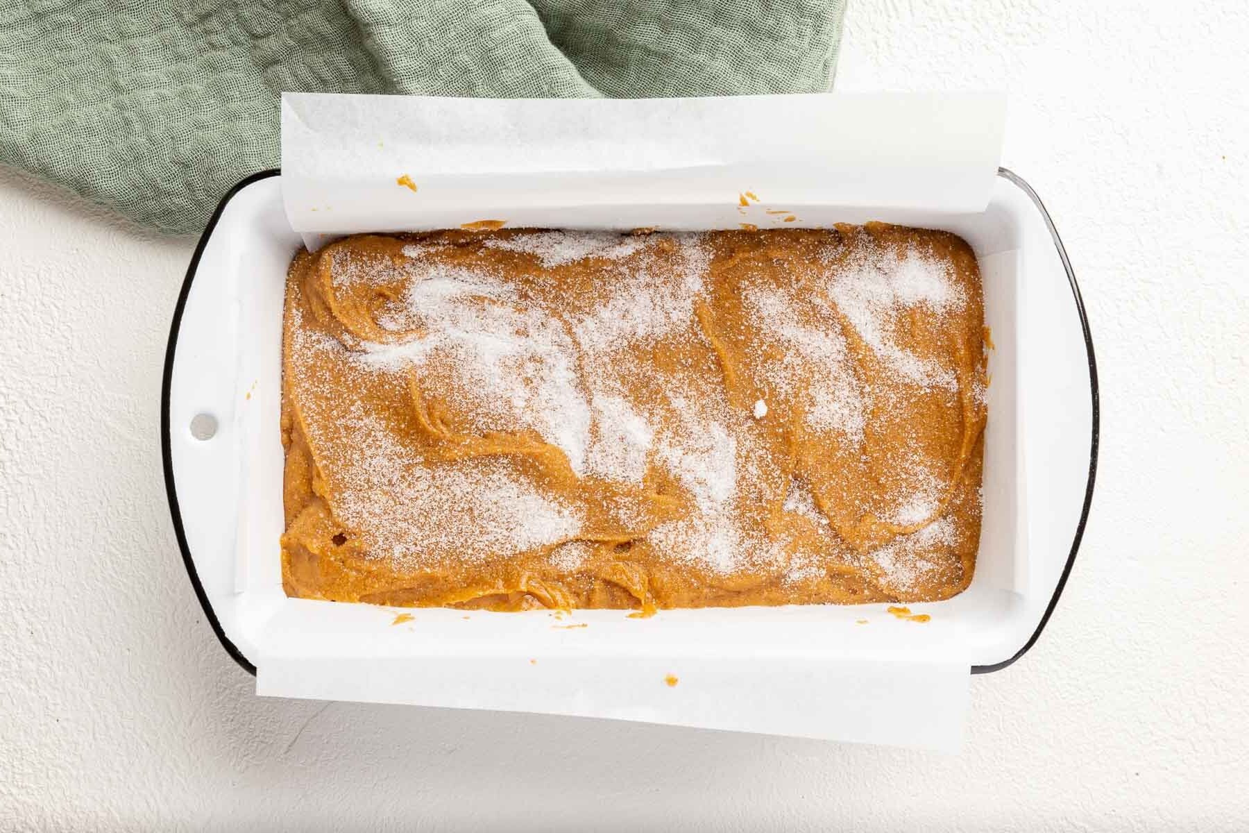 A white loaf pan lined with parchment paper and filled with gluten-free pumpkin bread batter and topped with granulated sugar.