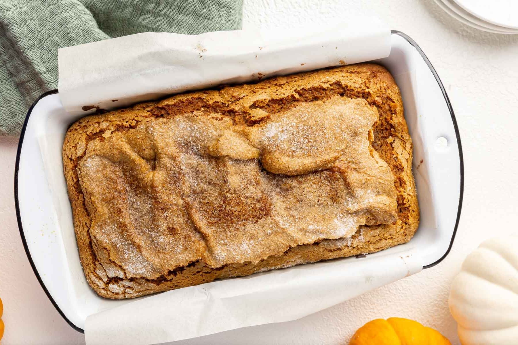 A loaf of gluten-free pumpkin bread in a white loaf pan with mini pumpkins to the side.