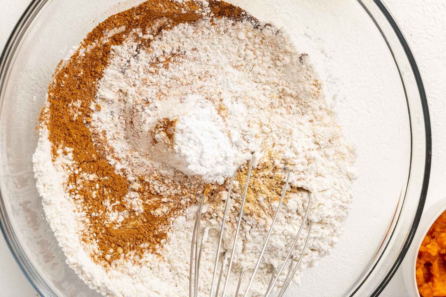 A glass mixing bowl with gluten-free flour, cinnamon, baking soda, baking powder, ginger, cloves, and nutmeg being mixed together with a whisk.